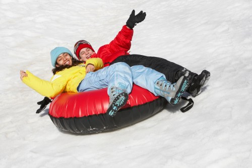 Two people in winter clothing joyfully sledding down a snowy hill on a red and black inner tube.