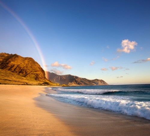 A serene beach scene with gentle waves, mountains in the background, and a rainbow arching across a clear blue sky.