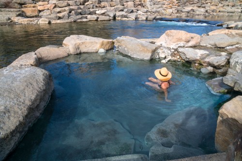 A person relaxes in a natural hot spring surrounded by rocks and water, wearing a wide-brimmed hat.