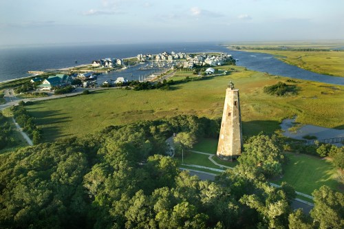 Aerial view of a lighthouse near a coastal town, surrounded by greenery and water, with houses in the background.