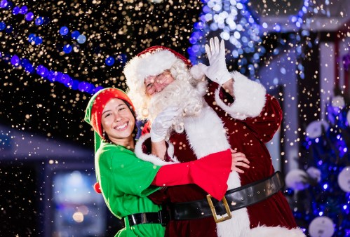 A smiling woman in an elf costume poses with Santa Claus, surrounded by festive lights and falling snow.