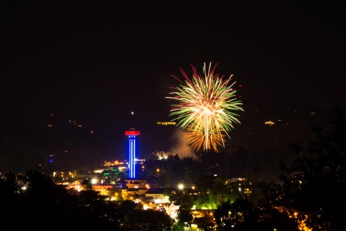 Colorful fireworks burst over a city skyline at night, with illuminated buildings and a tall tower in the foreground.