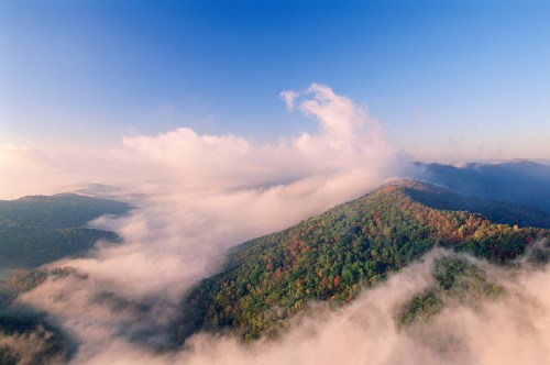 Aerial view of a lush green mountain landscape shrouded in mist under a clear blue sky.