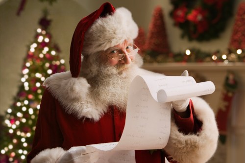 Santa Claus holds a long list, smiling, with a decorated Christmas tree in the background.