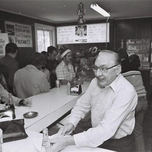 A man sits at a counter in a busy diner, surrounded by patrons and a menu board in a nostalgic black-and-white setting.