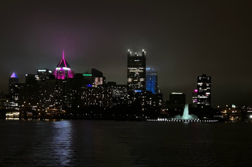 City skyline at night with colorful lights and a fountain, reflecting on the water under a foggy sky.