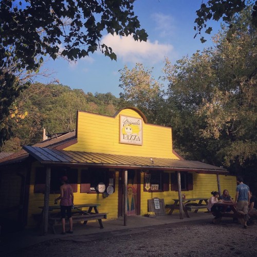 A bright yellow pizza shop surrounded by trees, with people sitting outside at picnic tables.