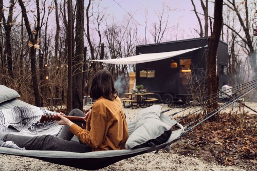 A person sits in a hammock playing guitar near a modern cabin in a wooded campsite at dusk.
