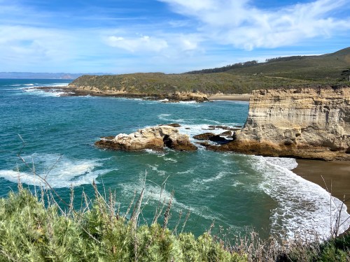 A scenic coastal view featuring cliffs, waves, and a sandy beach under a blue sky with scattered clouds.