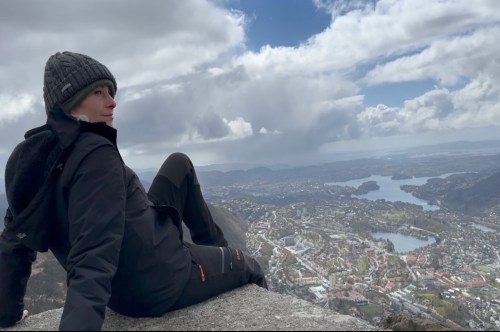 A person in a black jacket and beanie sits on a rocky ledge, overlooking a scenic landscape with mountains and a lake.