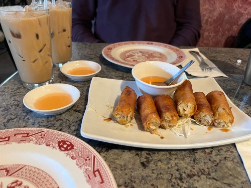 A plate of crispy spring rolls with dipping sauce, accompanied by two iced drinks on a restaurant table.
