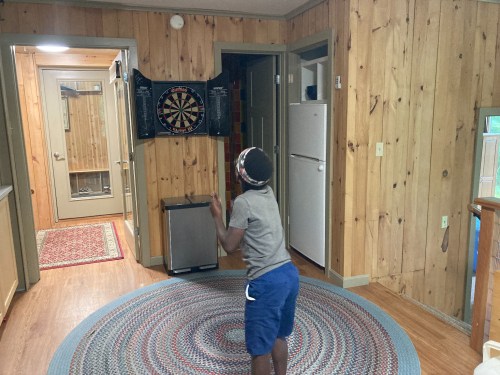 A child throws a dart at a dartboard in a wooden-paneled room with a circular rug and kitchen appliances visible.