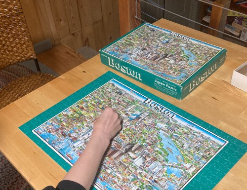 A person assembling a Boston jigsaw puzzle on a wooden table, with the puzzle box nearby.