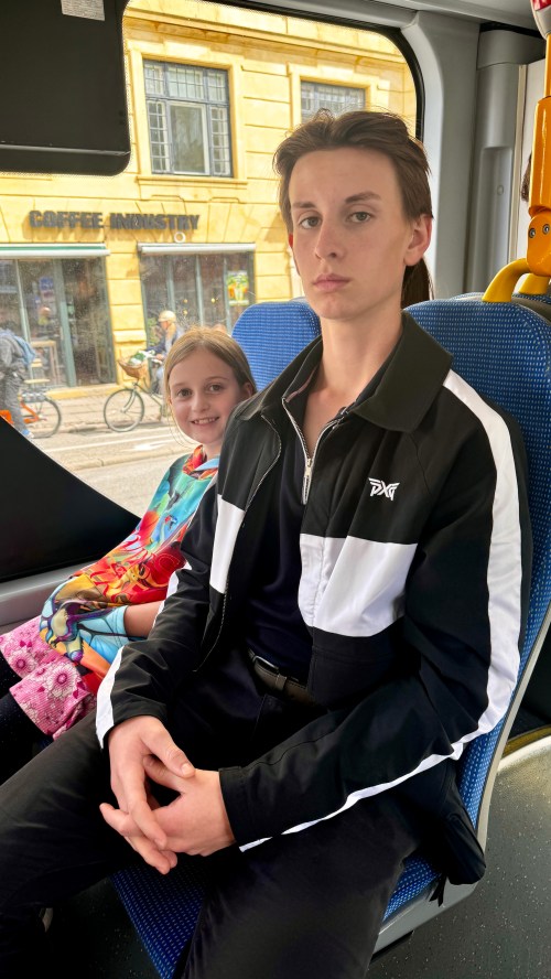 A young boy in a black and white jacket sits on a bus, with a girl in colorful clothing smiling behind him.