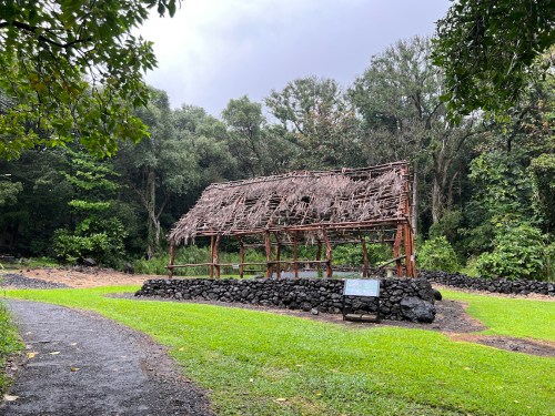 A rustic wooden structure with a thatched roof, surrounded by lush greenery and a gravel path.