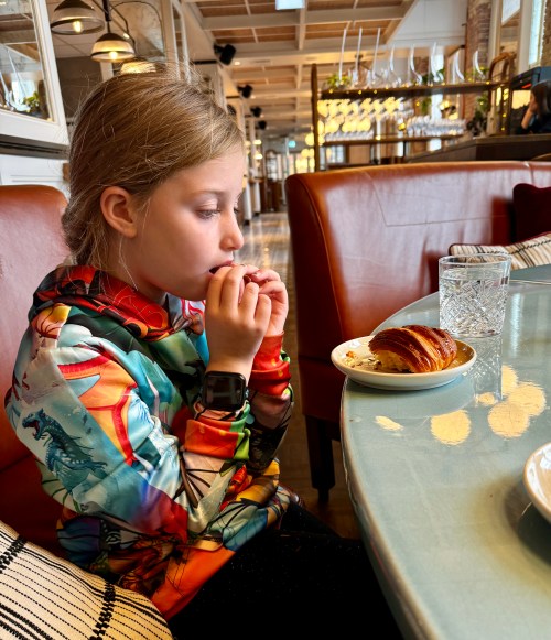 A young girl in a colorful sweatshirt sits at a table, thoughtfully eating a pastry with a glass of water nearby.