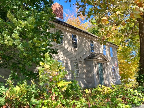 A historic house partially obscured by greenery, surrounded by colorful autumn foliage under a clear blue sky.