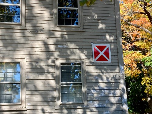 A weathered building with two windows and a red and white diagonal cross sign on the wall, surrounded by autumn foliage.