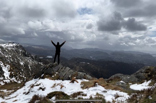 A person stands on a rocky outcrop with arms raised, overlooking a mountainous landscape under a cloudy sky.