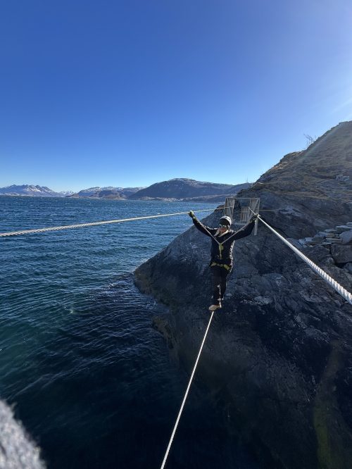 A person balances on a tightrope over water, surrounded by rocky terrain and mountains under a clear blue sky.