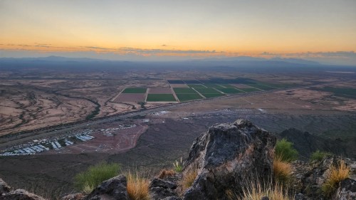 A panoramic view from a mountain peak overlooking fields and valleys at sunset, with rocky terrain in the foreground.