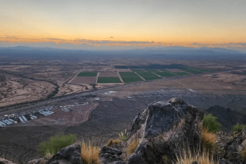A panoramic view from a mountain overlooking fields and a valley at sunset, with rocky terrain in the foreground.