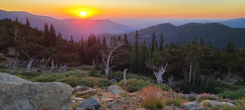 A vibrant sunset over mountains, with silhouetted trees and colorful skies, creating a serene natural landscape.