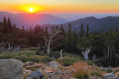 Sunset over a mountainous landscape, with vibrant colors illuminating the sky and trees in the foreground.