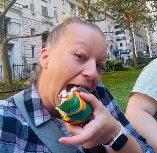 A person with a bun hairstyle is about to take a big bite of a colorful rainbow dessert in a park setting.