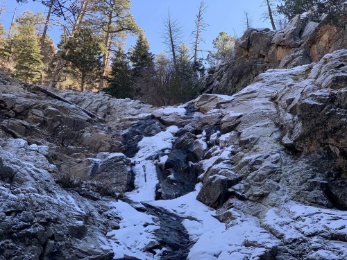 A rocky landscape with a small snow-covered waterfall, surrounded by trees and a clear blue sky.