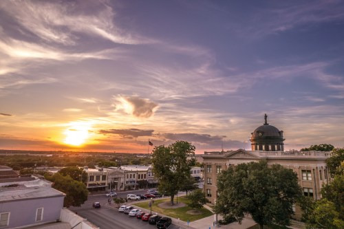 Sunset over a small town, featuring a historic building with a dome and trees, casting a warm glow in the sky.