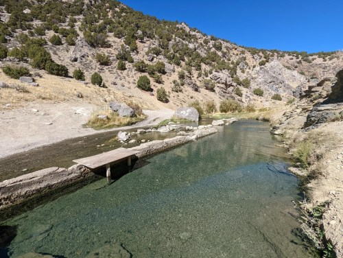 A clear stream flows through a rocky landscape, with a wooden bridge crossing over it under a blue sky.