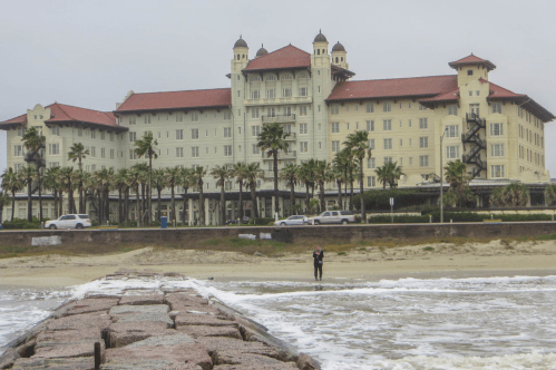 A beachfront view of a large hotel with palm trees, cloudy skies, and a person walking near the water.
