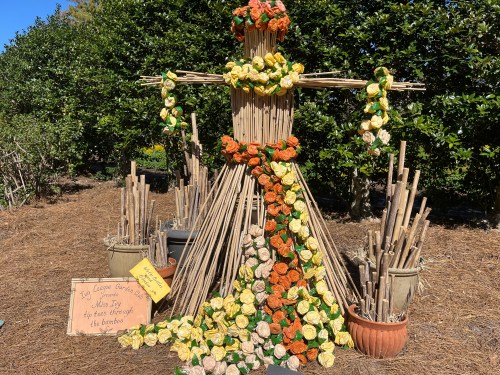 A decorative scarecrow made of bamboo and flowers, surrounded by potted plants in a garden setting.