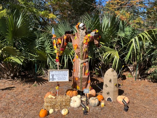 A scarecrow dressed in a brown robe, adorned with autumn leaves, surrounded by pumpkins and a beehive in a garden setting.