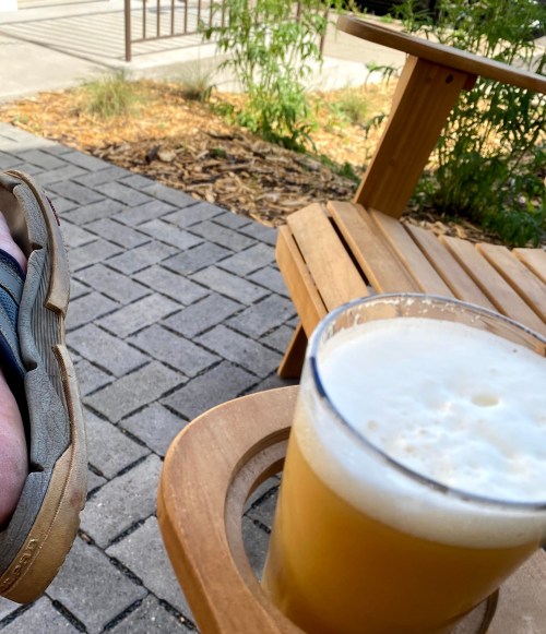 A close-up of a beer in a glass on a wooden chair, with a foot resting nearby on a stone patio.
