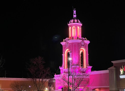 A brightly lit tower with pink lights at night, featuring decorative elements and a statue on top.