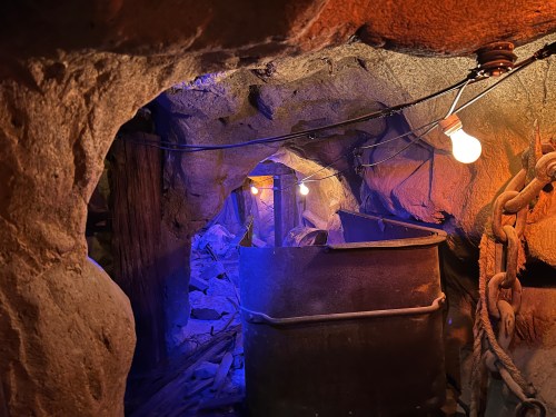 Dimly lit mine tunnel with a metal cart, illuminated by hanging light bulbs and surrounded by rocky walls.