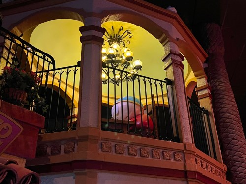 A colorful, whimsical balcony with a chandelier and a large, cartoonish character peeking over the railing.