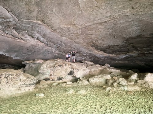 Two people stand on a rocky surface inside a large cave, with high ceilings and textured stone walls.