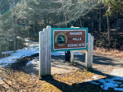 Sign for Wagner Falls Scenic Site, surrounded by trees and snow, indicating a nature area for visitors.