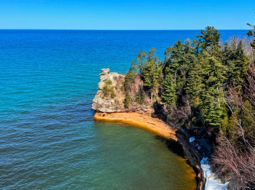 A rocky shoreline with lush green trees, overlooking a calm blue lake under a clear sky.