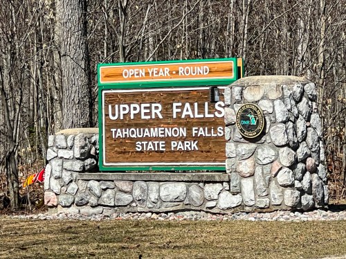Sign for Upper Falls at Tahquamenon Falls State Park, indicating it's open year-round, with a stone base and wooded background.