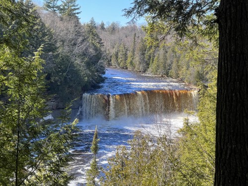 A scenic view of a waterfall surrounded by lush green trees and a clear blue sky. Water cascades over the rocky edge.