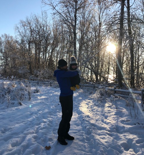 A person holds a child in winter clothing, surrounded by snow-covered trees and a bright sun in the background.