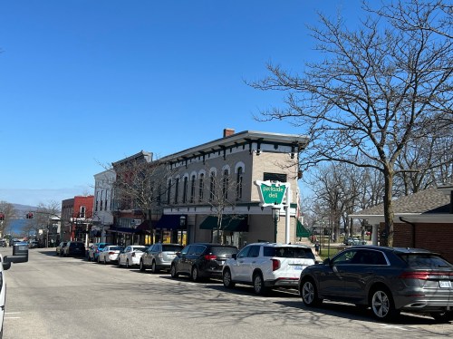 A sunny street view of a small town with parked cars and historic buildings, including a deli sign.
