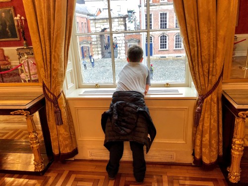 A child leans against a window, looking out at a cobblestone courtyard and historic buildings.