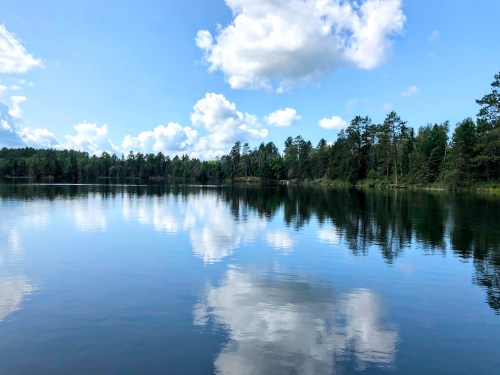 A serene lake reflecting fluffy clouds and trees under a bright blue sky.