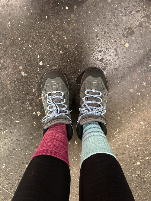 A pair of hiking shoes worn with mismatched socks: one pink and one light blue, on a textured floor.