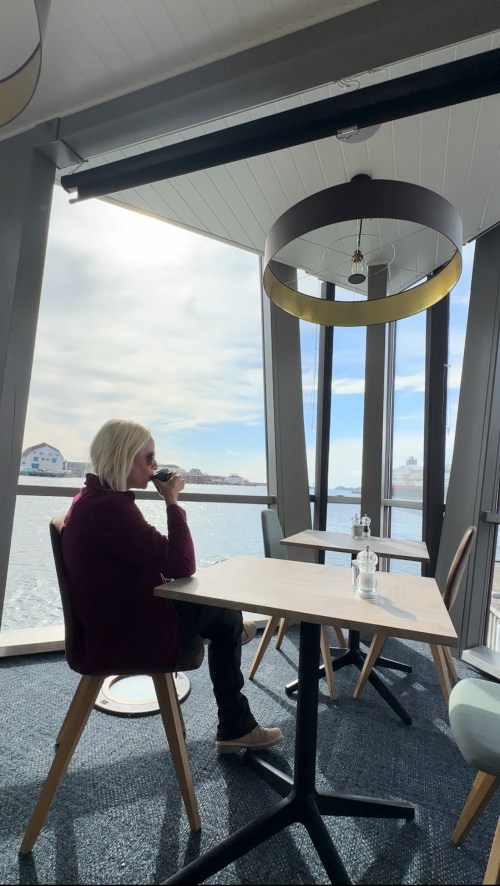 A person with blonde hair sits at a table by a large window, sipping a drink with a view of the water outside.